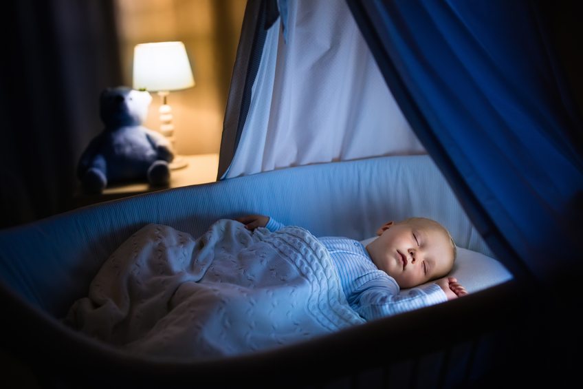 Adorable baby sleeping in blue bassinet with canopy at night. Little boy in pajamas taking a nap in dark room with crib, lamp and toy bear. Bed time for kids. Bedroom and nursery interior.