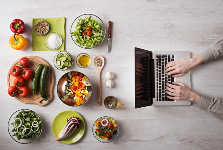 Man in the kitchen searching for recipes on his laptop with food ingredients and fresh vegetables on the left, top view