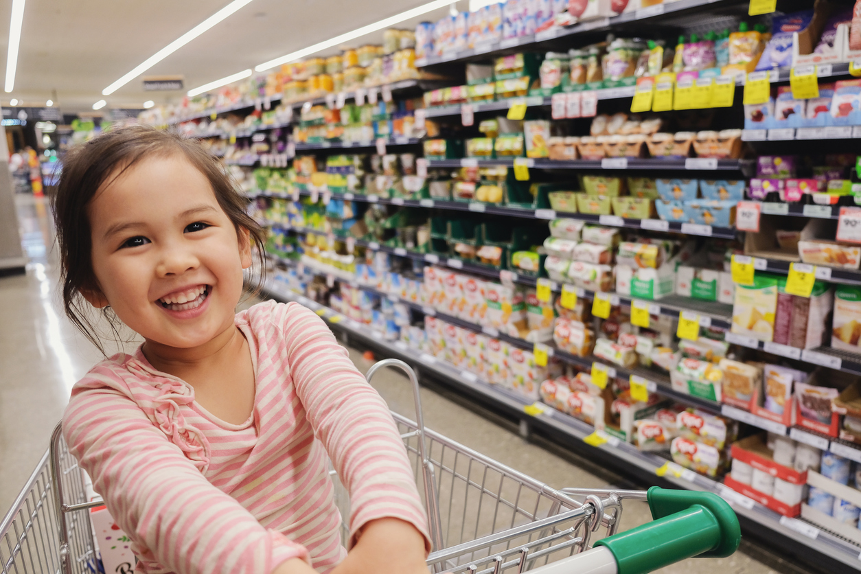Happy little multiethnic girl sitting in a trolley, shopping cart at supermarket, grocery store